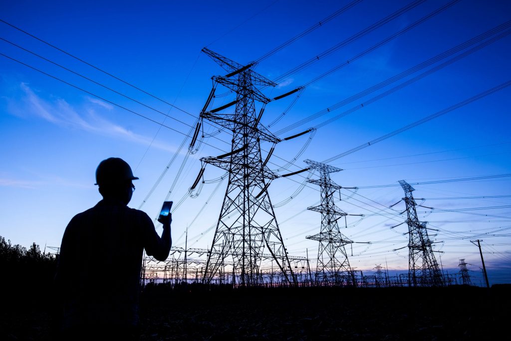 setor elétrico - Homem com um celular em frente a uma torre de energia elétrica sob o céu de fim de tarde.